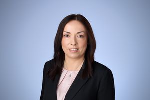 Professional headshot of a dark-haired woman in a black blazer and pink blouse against a blue background.