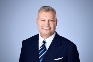 Professional headshot of a man with gray hair wearing a navy suit and striped tie against a blue background.