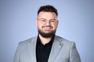Smiling young man with beard and glasses wearing a light gray blazer and black shirt against a blue background.