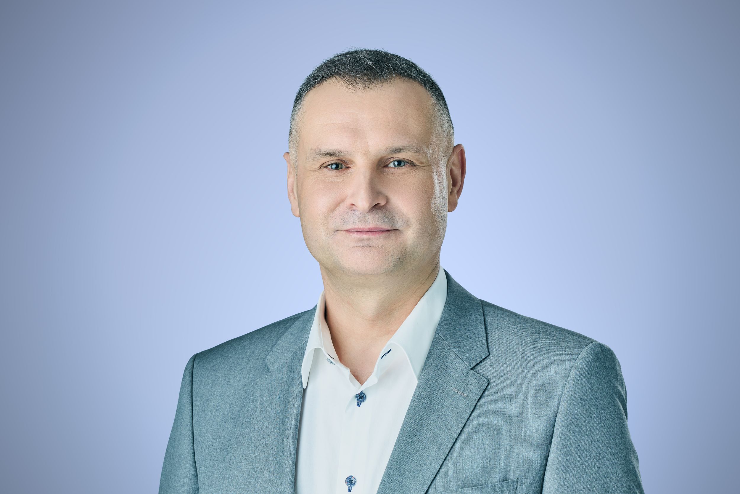 Professional headshot of a middle-aged man in a gray blazer smiling confidently against a light blue background.