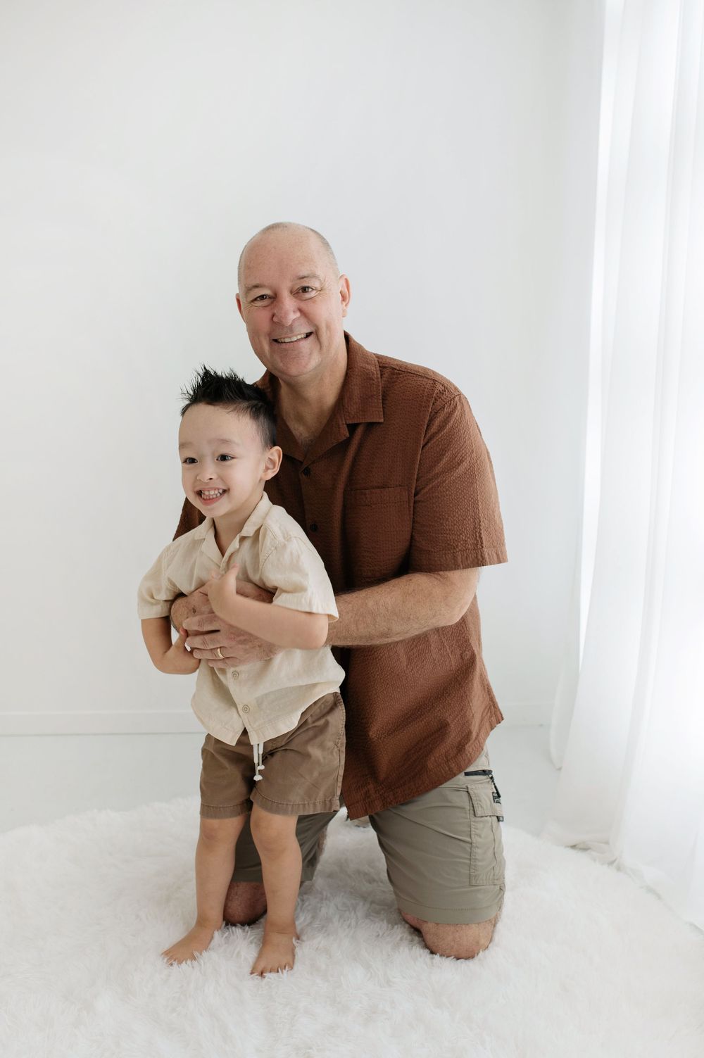 Smiling bald grandfather kneeling behind happy toddler boy, both wearing matching khaki outfits on white fluffy rug.
