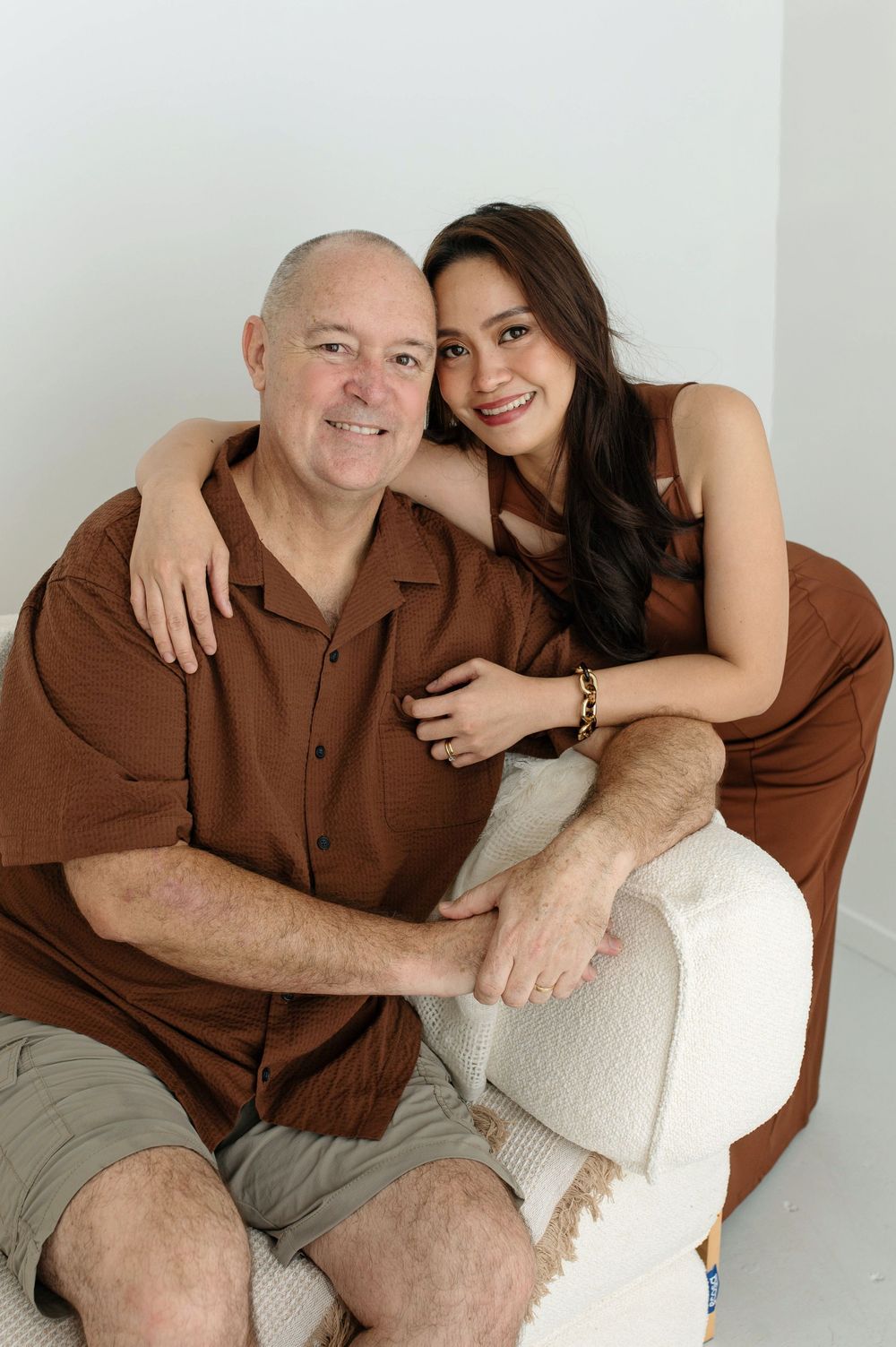 A smiling older man and young woman in matching brown outfits pose together on a white chair in a bright studio.