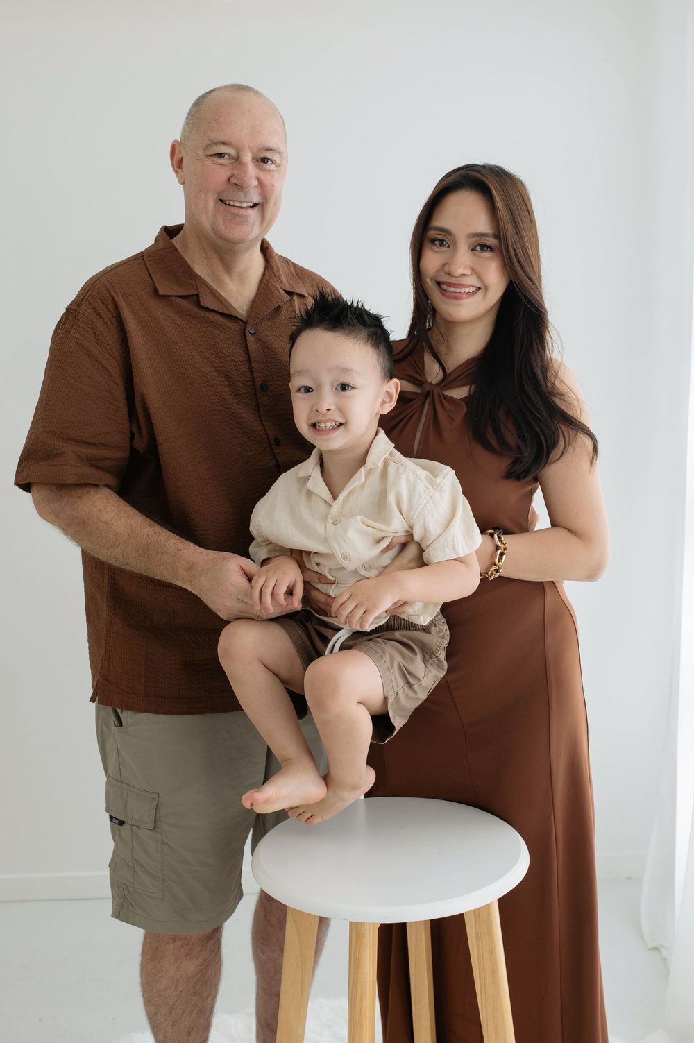A smiling family of three poses for a portrait, with parents in brown outfits and a young boy seated on a white stool.