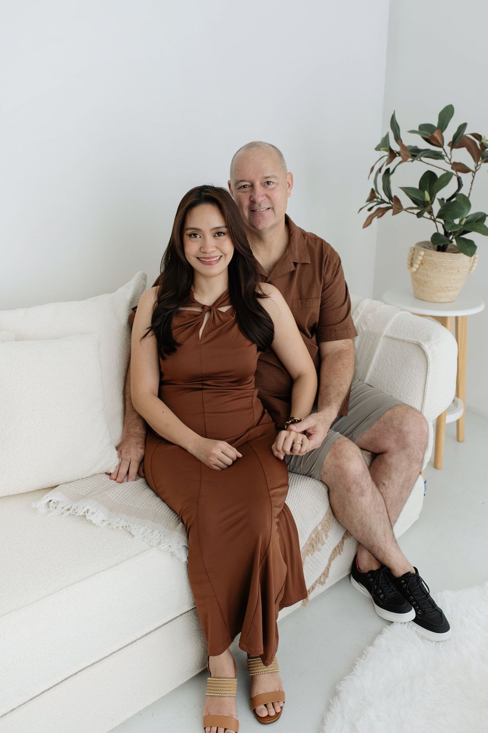 A couple in matching brown outfits sitting together on a white couch in a bright, minimalist studio setting.