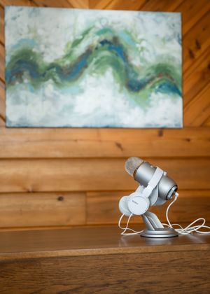 Silver USB microphone on wooden desk with abstract green and blue wave painting on wood panel wall in background.