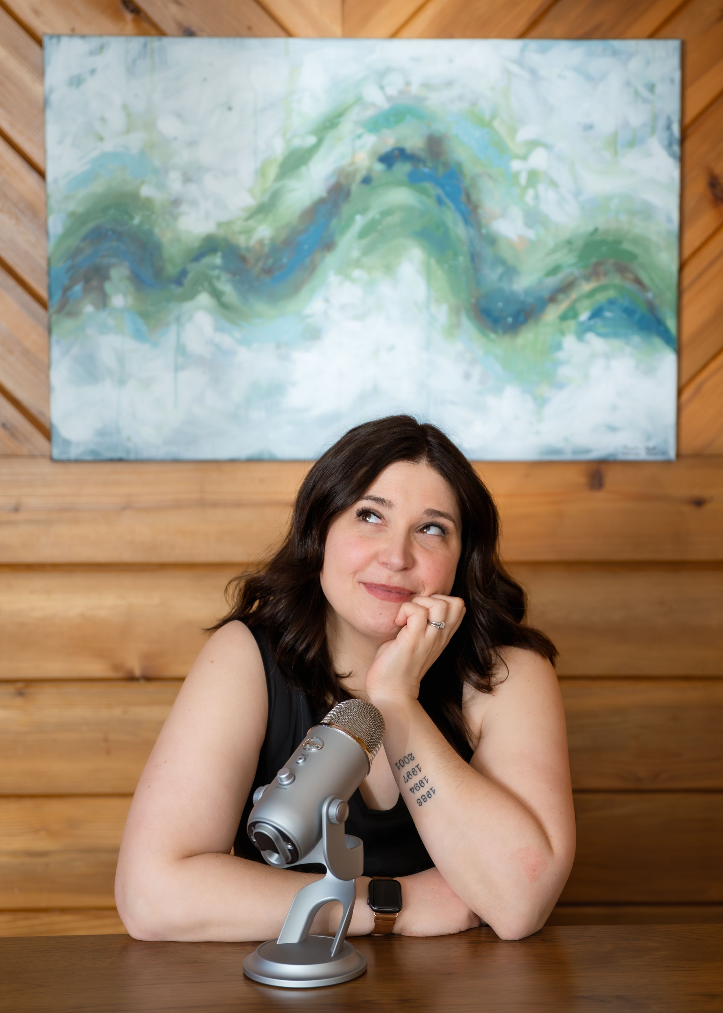 Woman sitting at a desk with a microphone, looking up thoughtfully, with abstract green and blue wall art behind her.