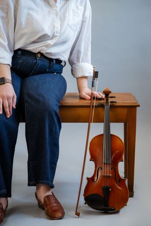 Person in white shirt and jeans sitting on wooden bench holding violin bow beside a classic violin.