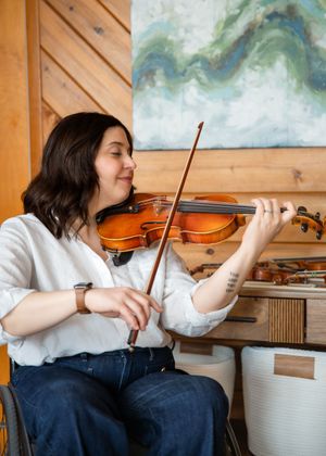 Woman in wheelchair playing violin in a cozy wooden room with abstract art painting on the wall behind her.