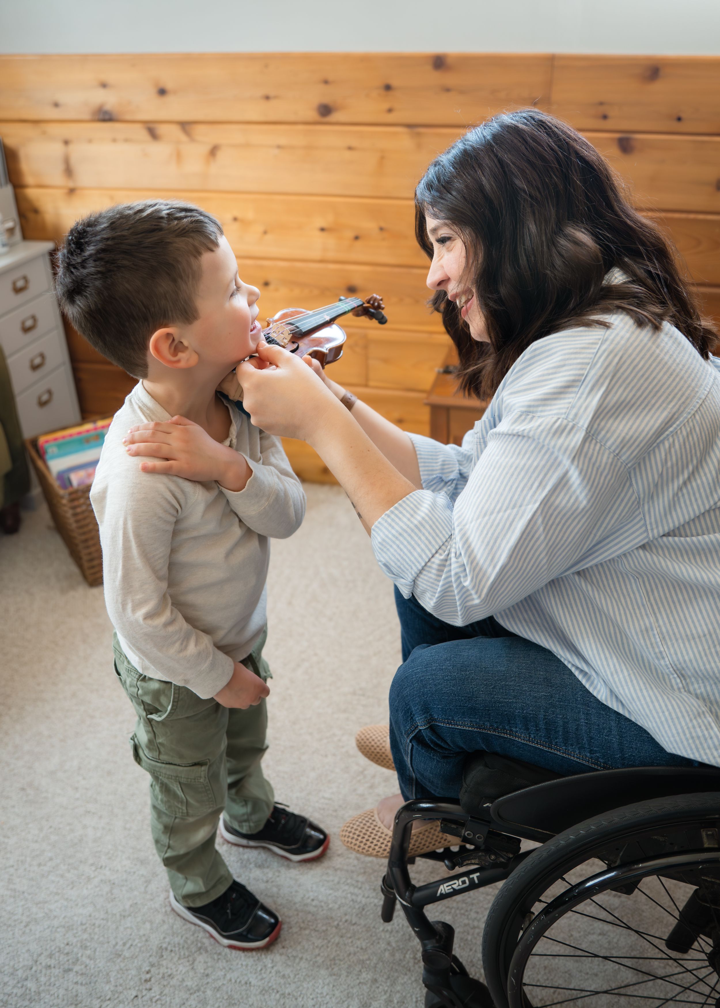Woman in wheelchair teaches young boy violin in a warm wooden room.