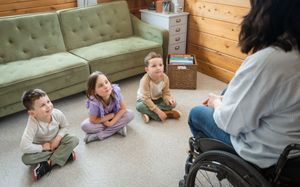 A teacher in a wheelchair engages three young children sitting on the floor in a cozy living room setting.