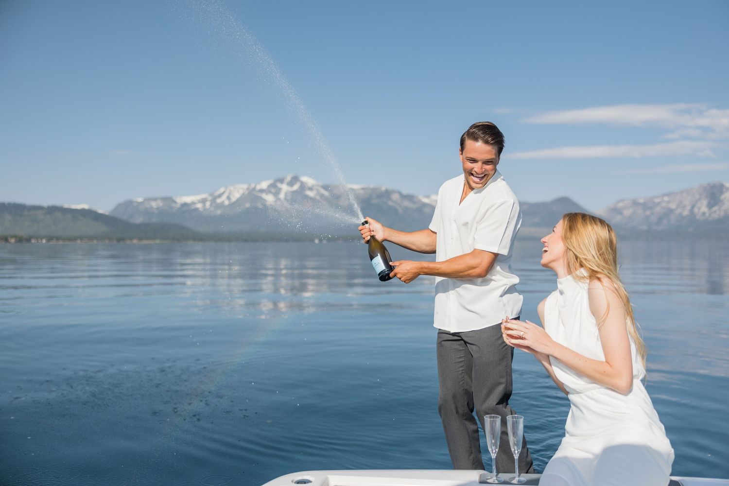 A white wedding cake on a boat with a blurred couple kissing in the background on a sunny day.