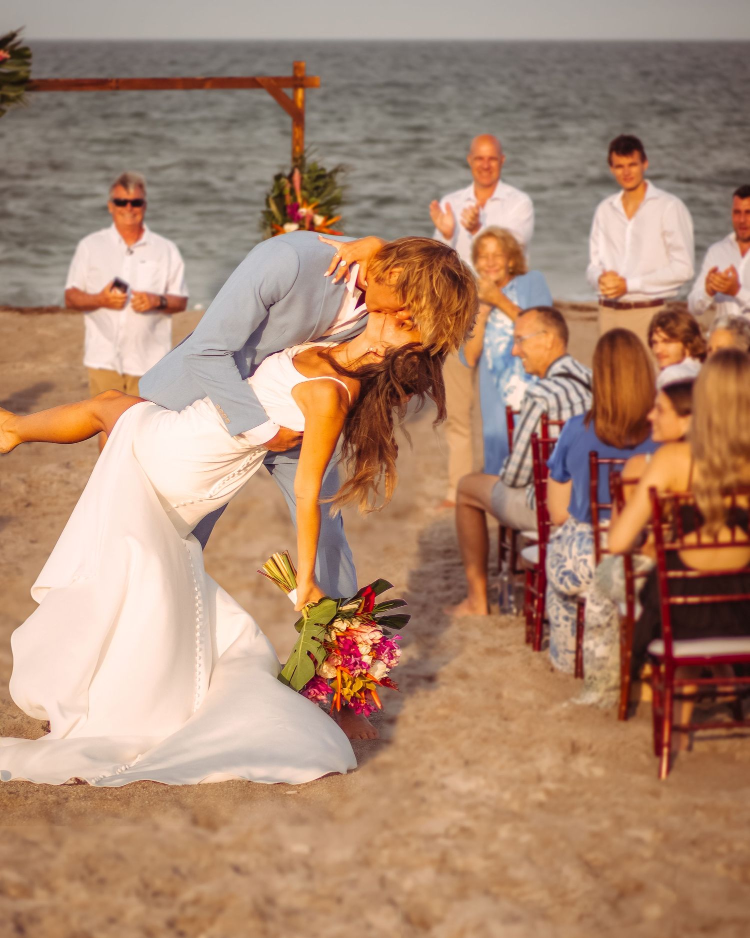 Bride and groom share a romantic dip kiss on a beach wedding ceremony with guests applauding in the background.