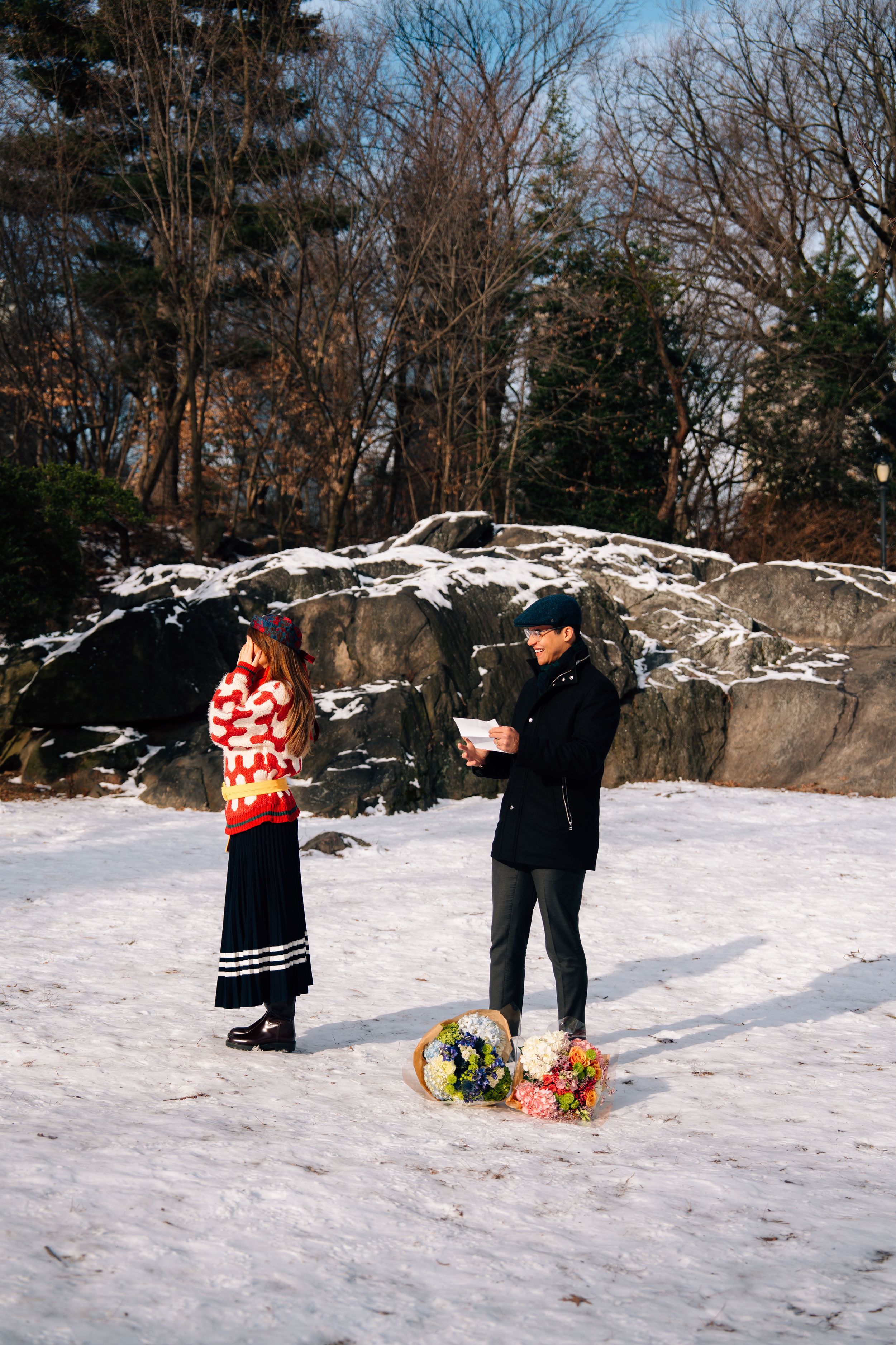 Two people stand on a snowy landscape in winter, with colorful flowers on the ground and rocky trees in the background.