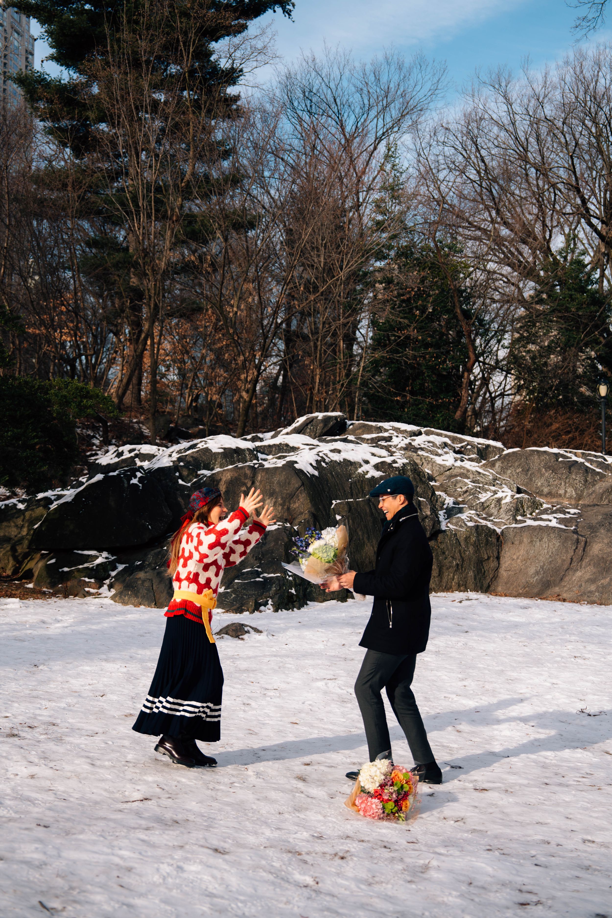 Couple exchanging flowers in a snowy park during winter, surrounded by bare trees and large rocks.