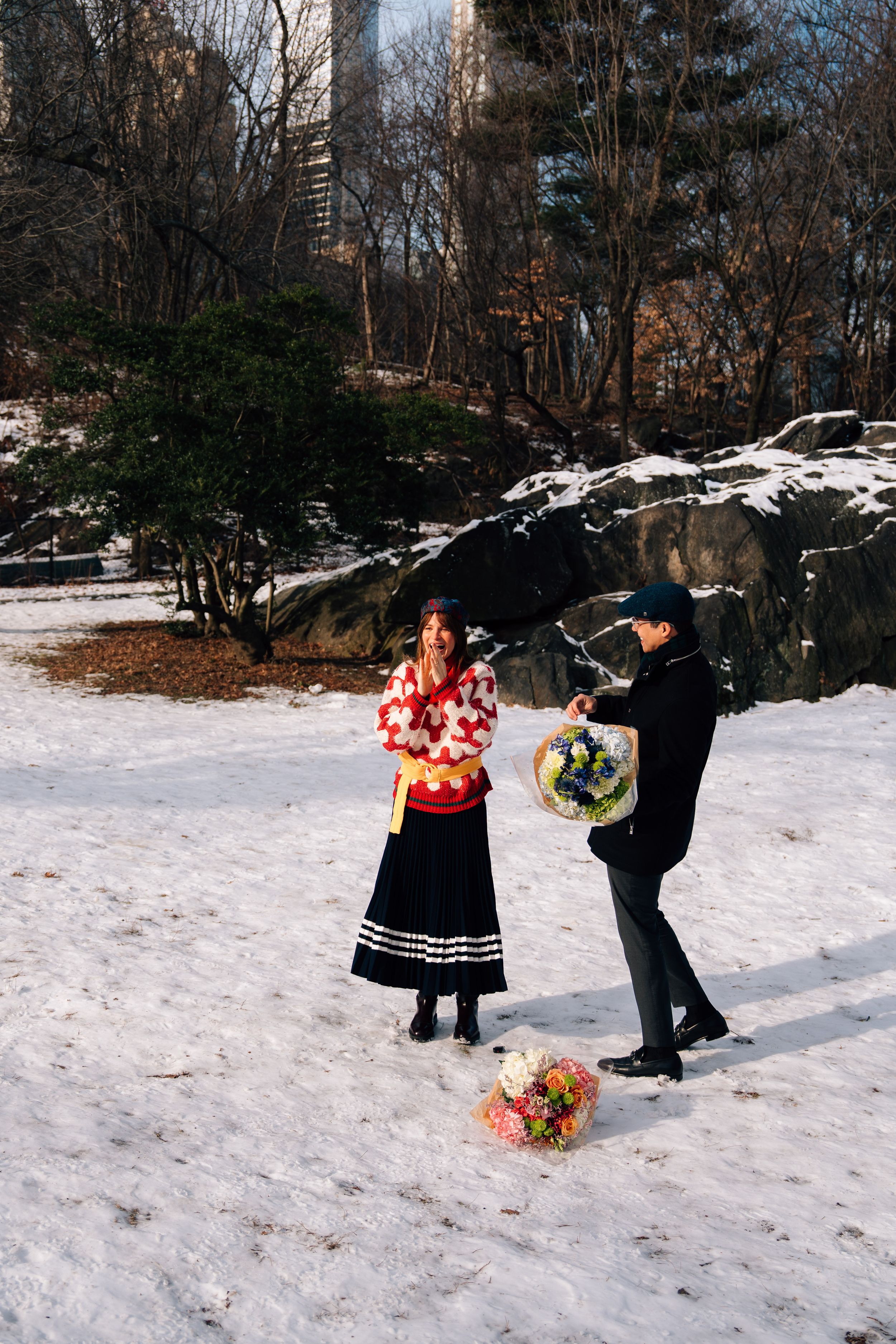 Couple exchanging flowers in snowy Central Park winter scene with rocks and trees in background.