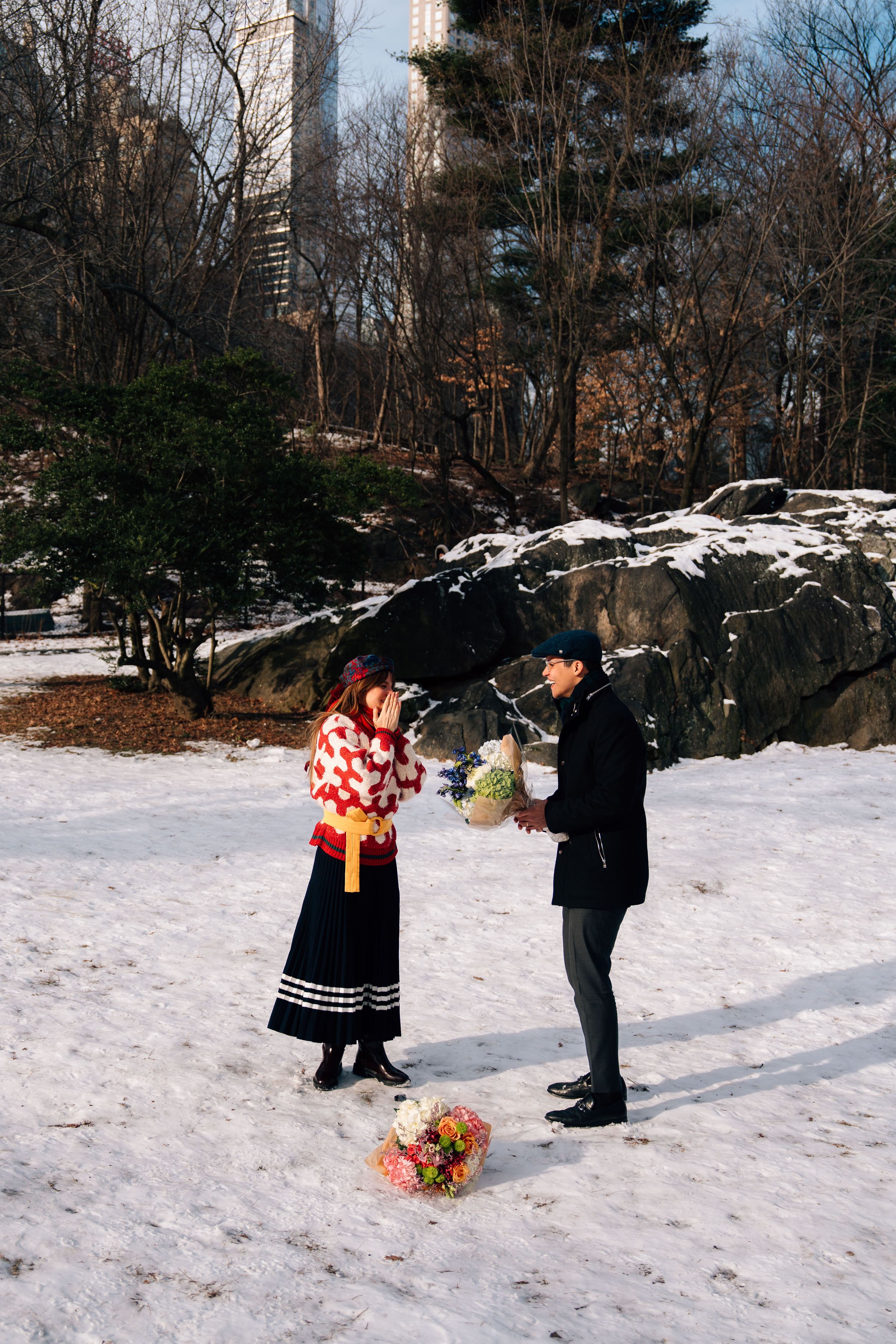 Couple exchanging flowers in snowy Central Park, NYC, with rocks and bare trees in winter backdrop.