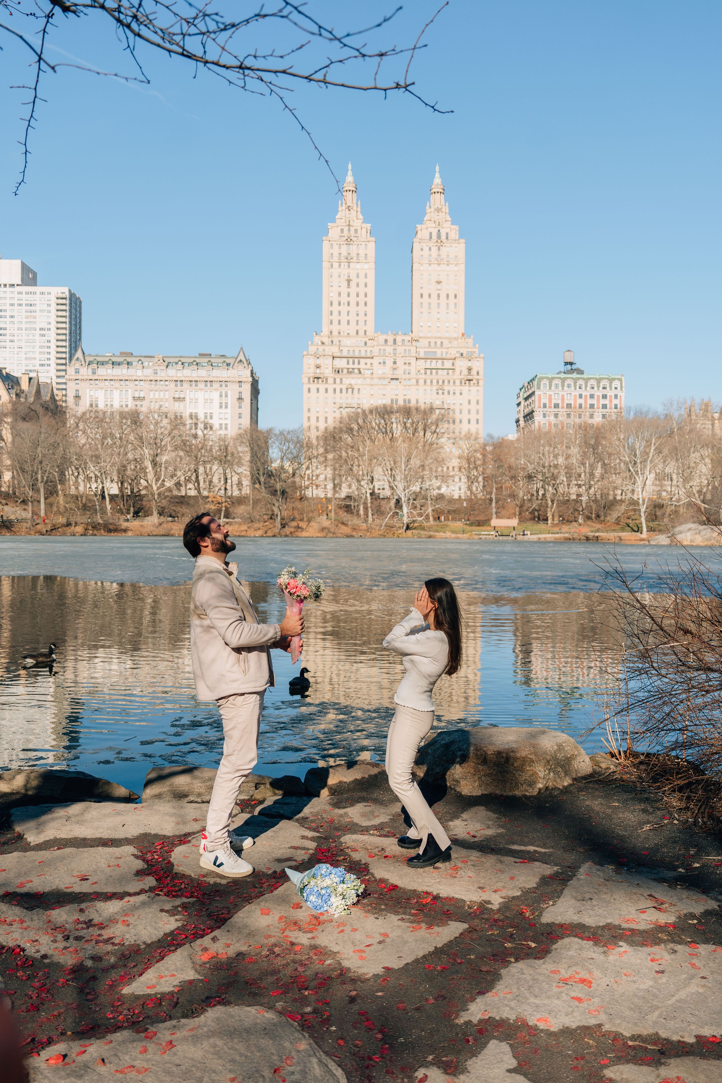 Couple celebrating a romantic proposal by a lake in Central Park, NYC, with iconic twin towers in the background on a clear day.