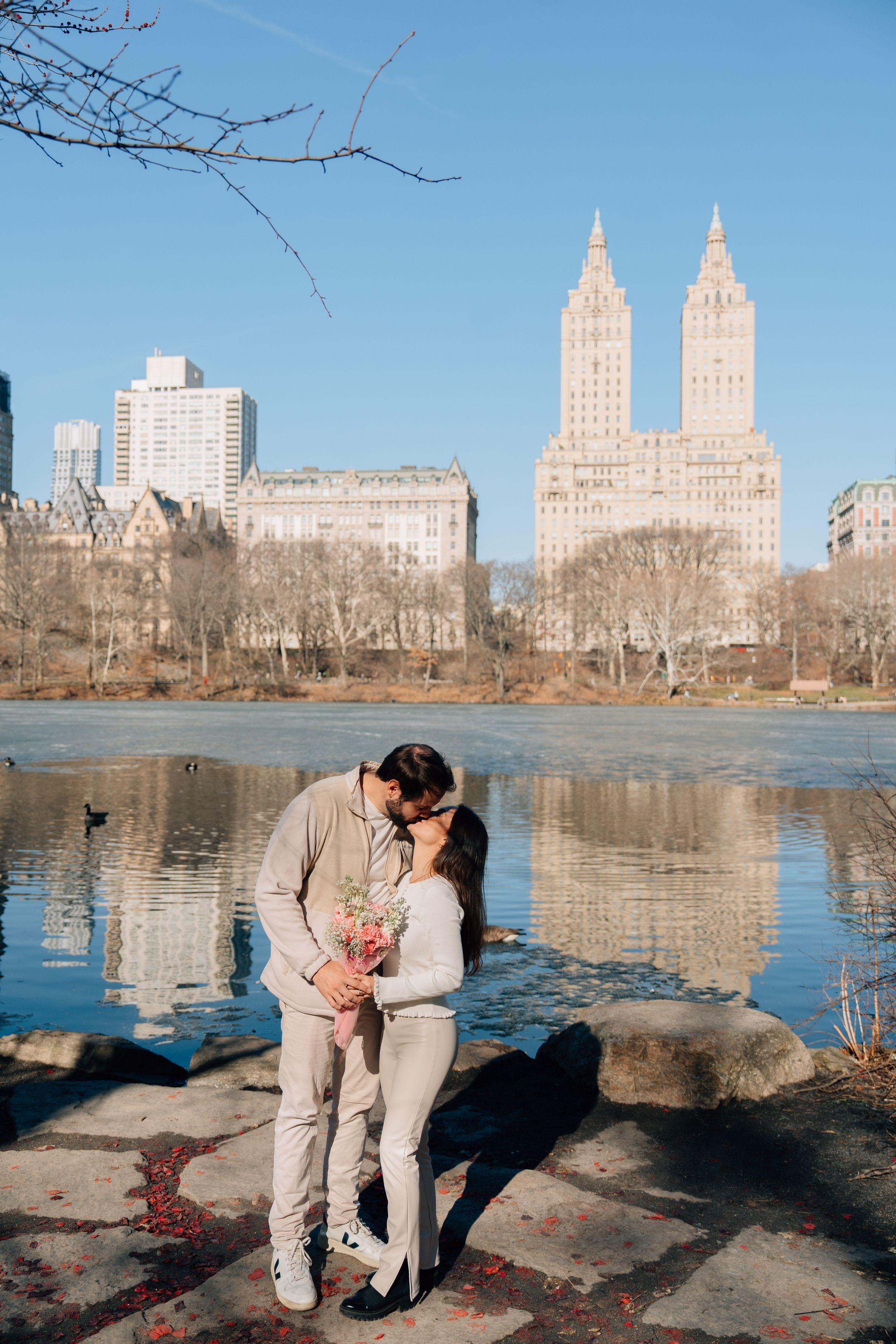 Couple kissing by Central Park lake with iconic San Remo building reflecting in water on a clear sunny day.