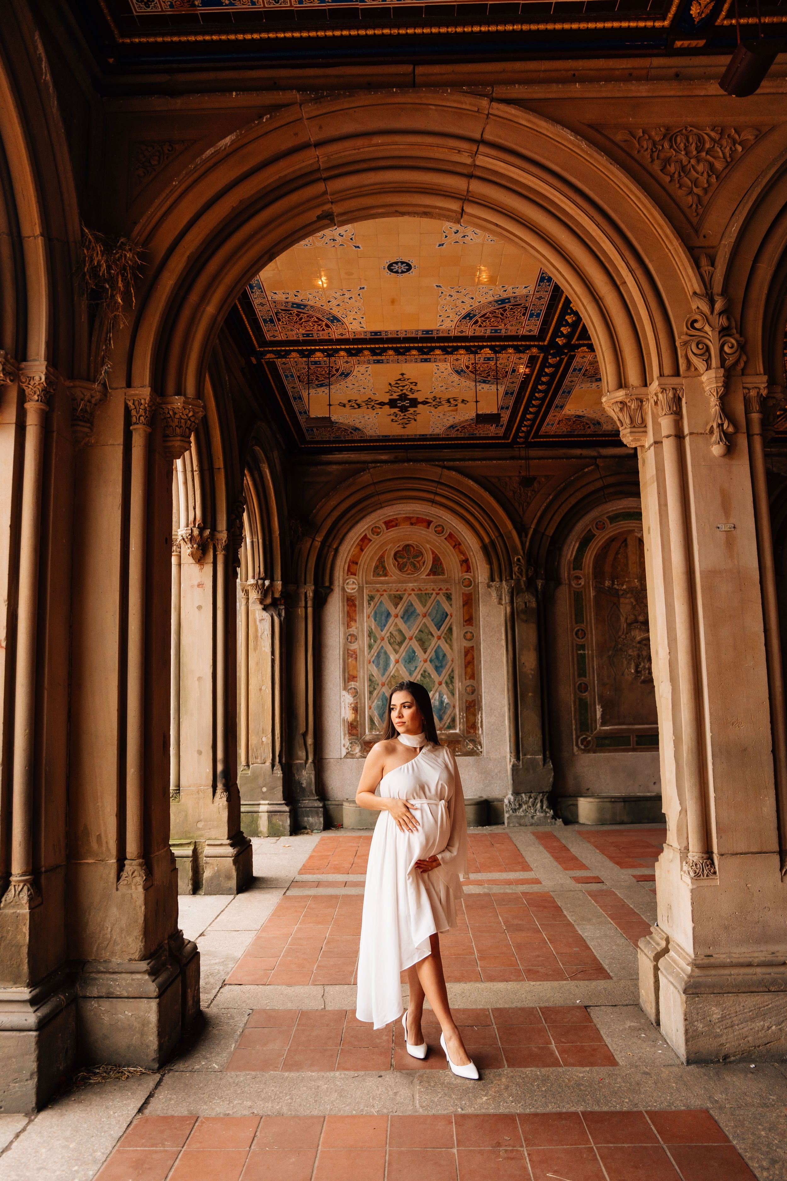 A bride in white dress poses in an ornate archway corridor with terracotta floors.