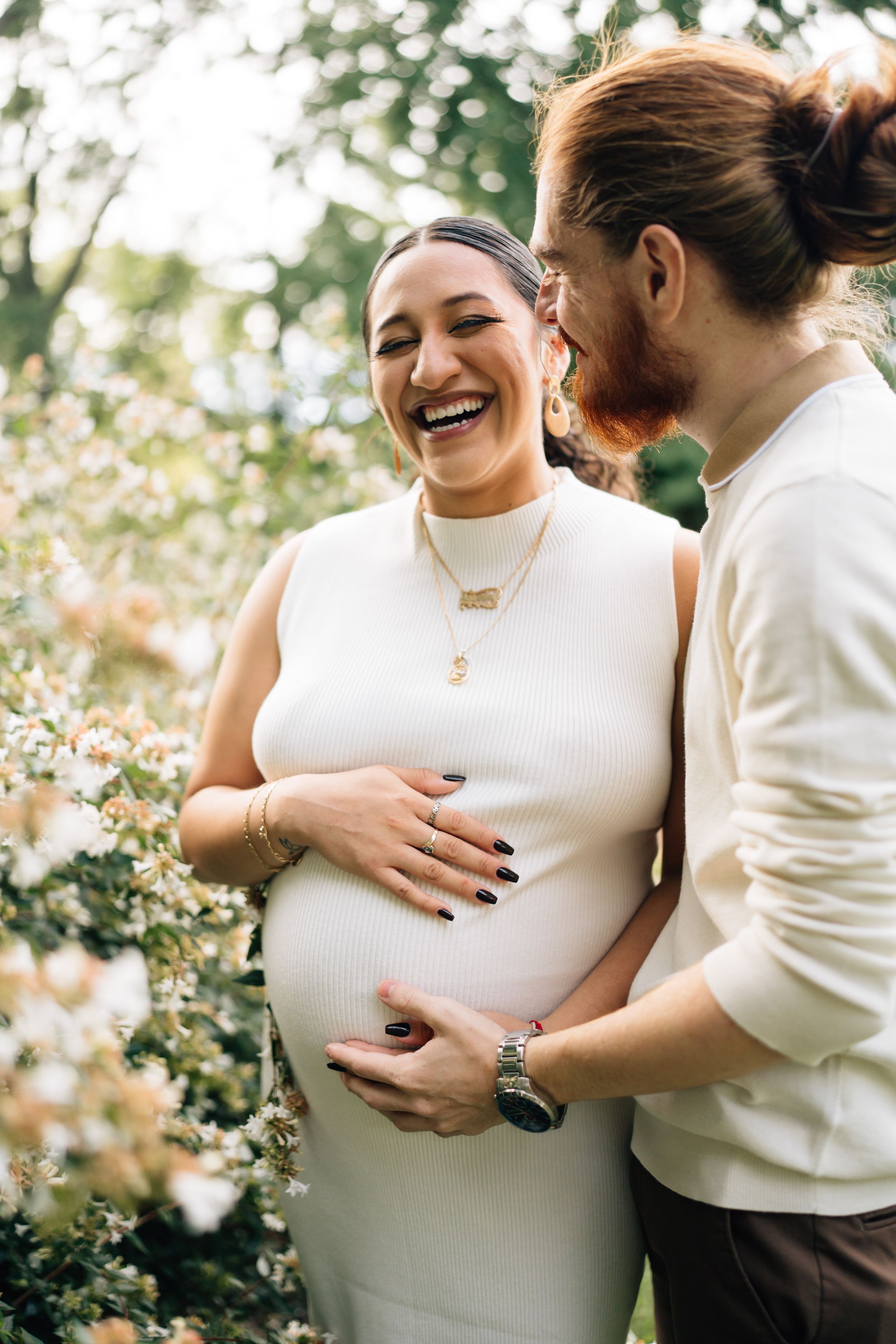 A candid maternity moment captured between two people in white sweaters among blooming flowers.