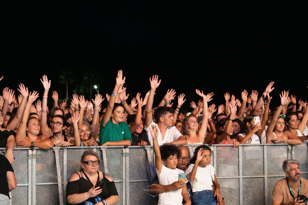 Excited concert crowd at night with hands raised, standing behind metal barriers at an outdoor music festival.