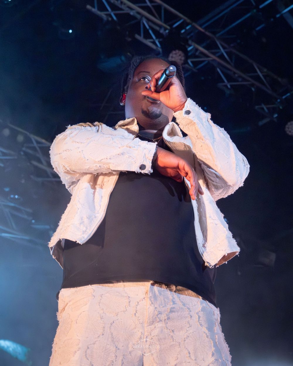 Performer in white lace jacket sings into microphone on stage under blue concert lights.