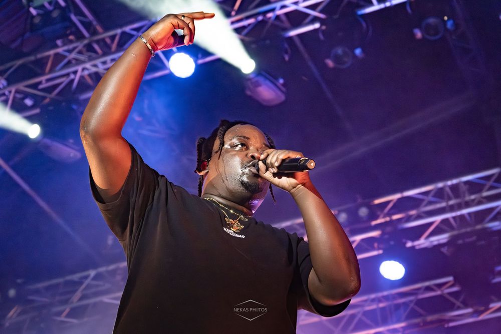 Male rapper performing on stage, pointing finger up, holding microphone under colorful concert lights.