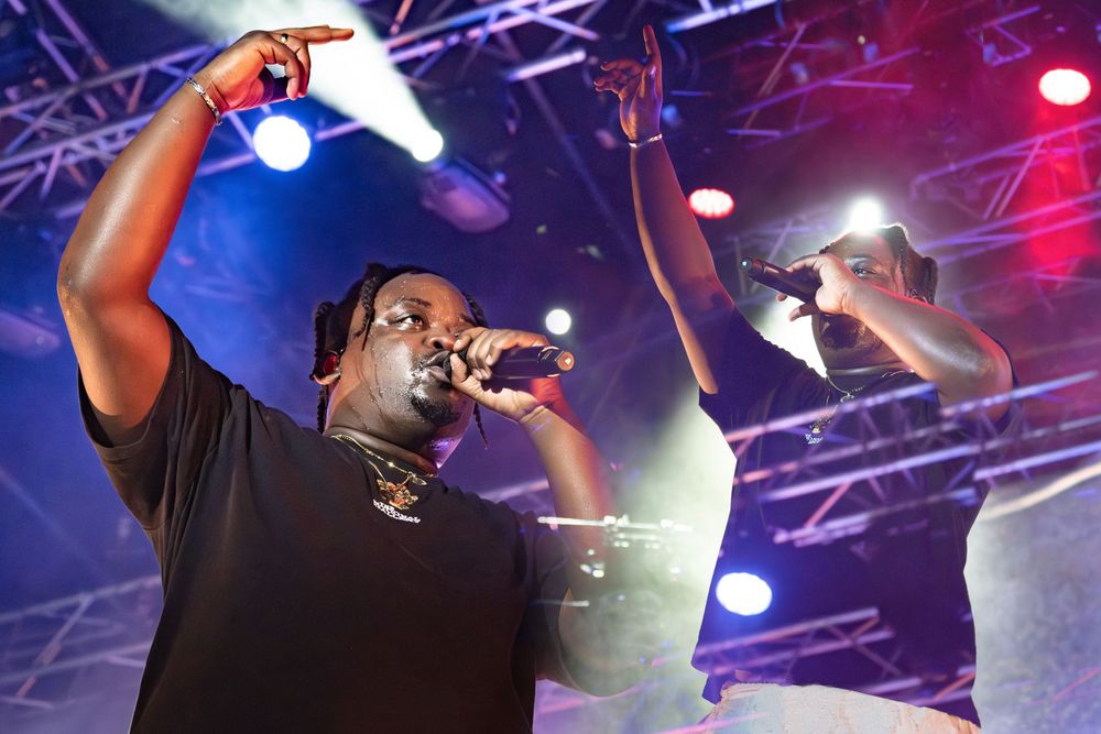 Two hip-hop artists performing on stage under colorful lights, one pointing up while holding a microphone.