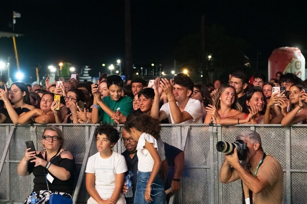 Excited crowd at an outdoor night concert, fans taking photos with smartphones behind a metal barrier.