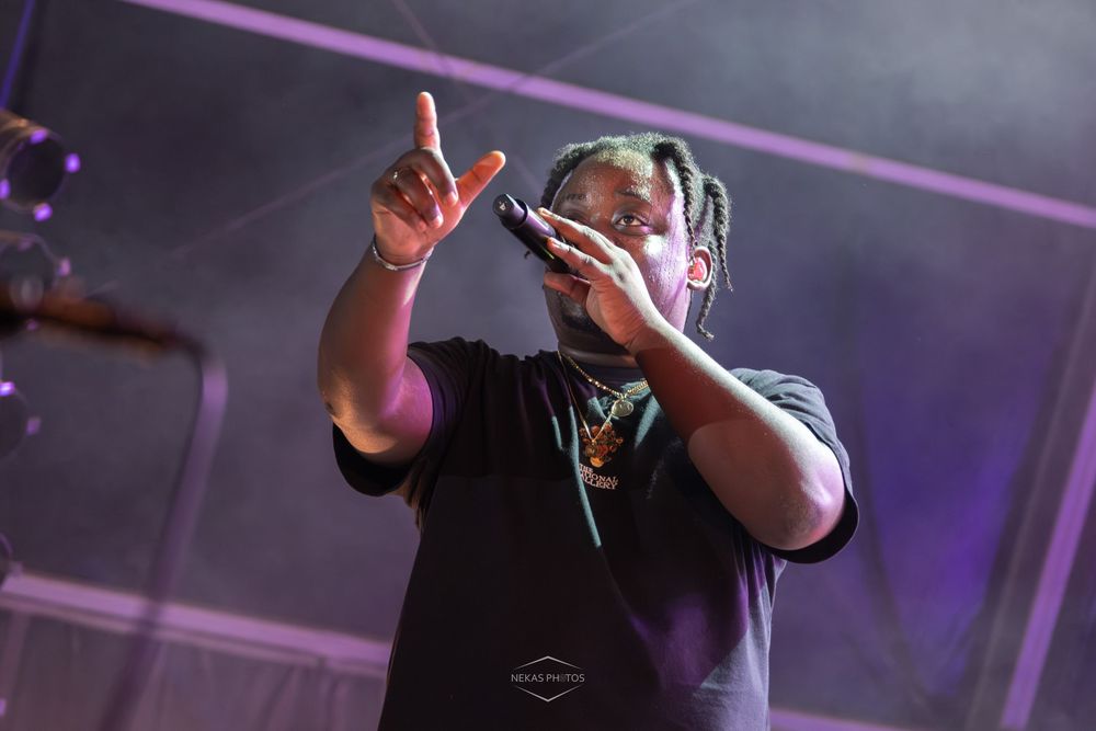 A rapper performing on stage, holding a microphone with one hand raised, wearing a black t-shirt under purple stage lights.