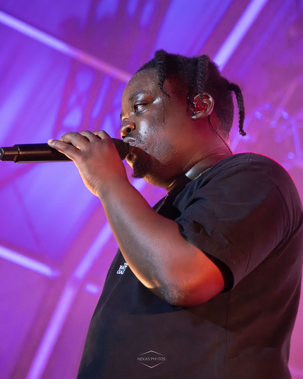 A male performer with braids sings into a microphone on a purple-lit stage wearing a black t-shirt.