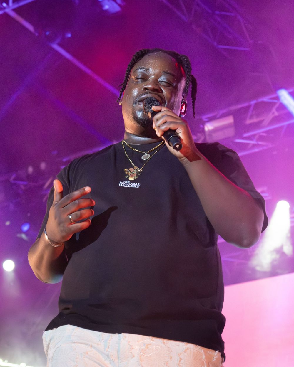 A performer sings into a microphone on stage wearing a black t-shirt and gold chain necklace under purple stage lights.