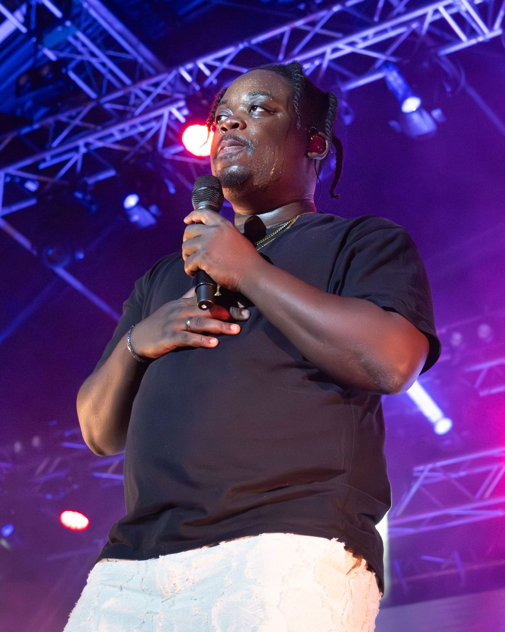 A performer in a black t-shirt holds a microphone on a concert stage with colorful lighting.
