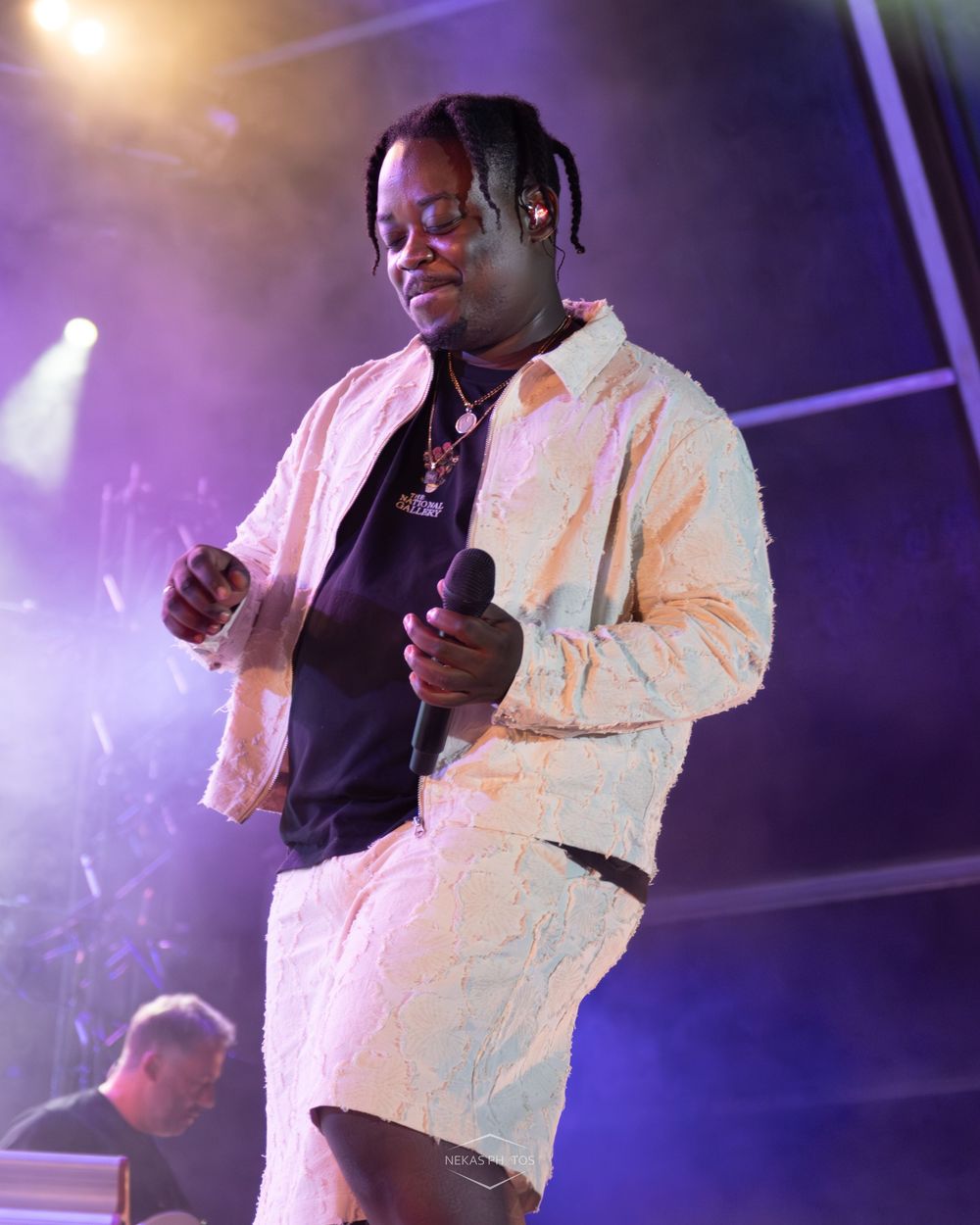 Performer in white textured outfit holds microphone on stage with purple lighting during live music event.