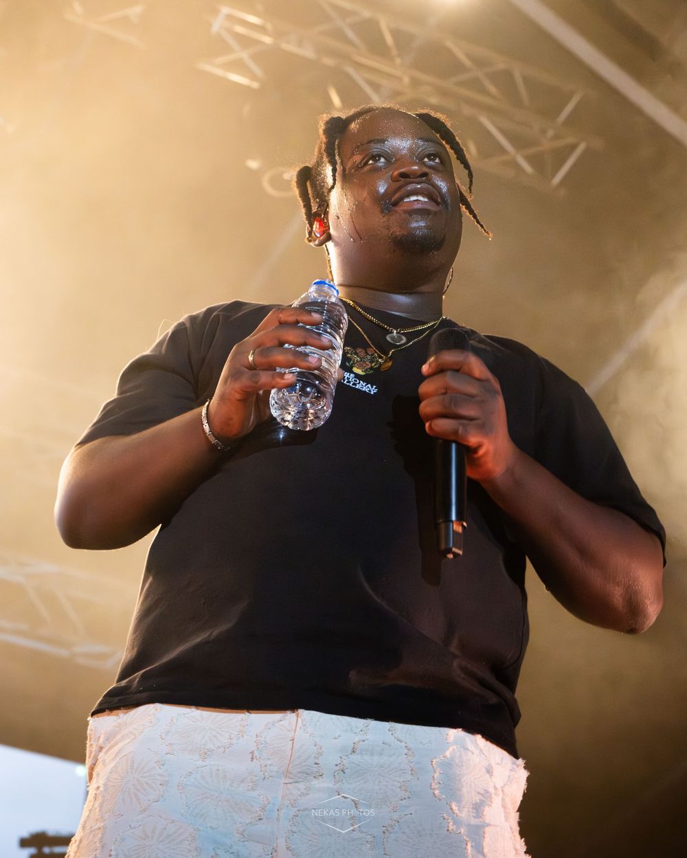 A performer on stage holds a microphone and water bottle, wearing a black t-shirt and white pants under stage lighting.