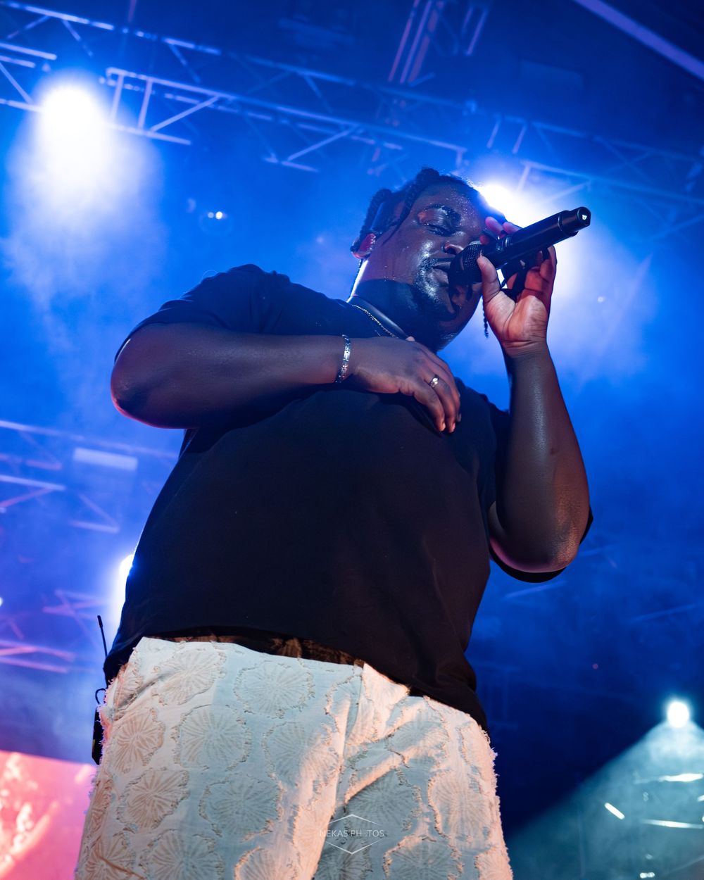 A Black male performer sings into a microphone on a blue-lit concert stage wearing a black shirt and white pants.