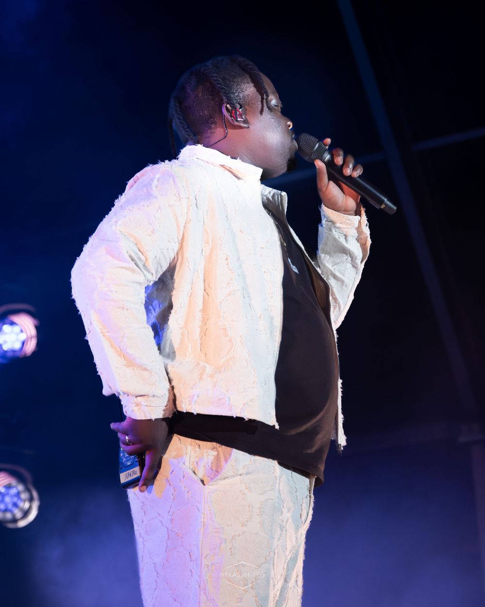 Performer in white lace outfit singing into microphone on stage with colorful lights at live concert.