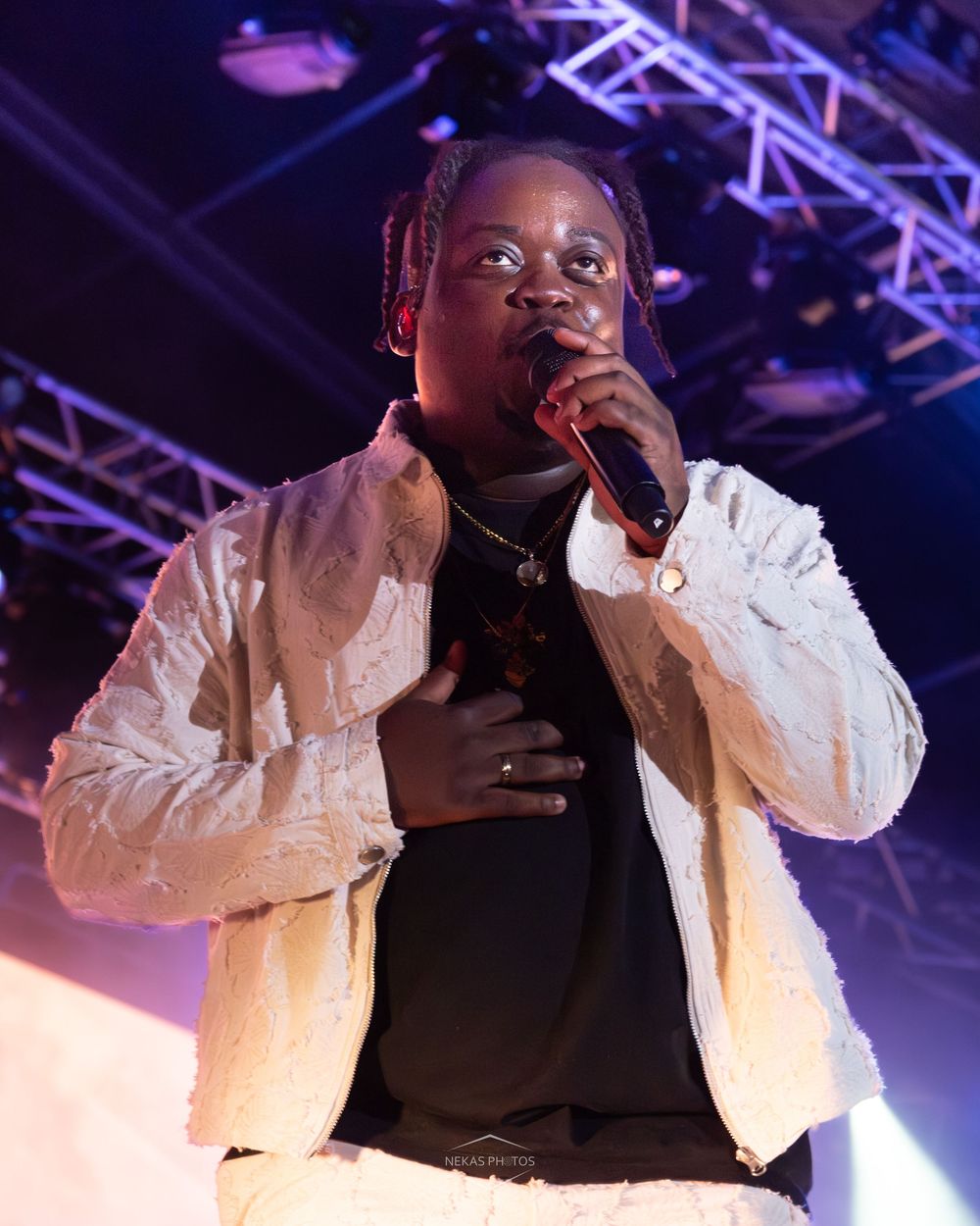 Young male rapper performing on stage, holding microphone, wearing white fuzzy jacket over black shirt with braided hair.