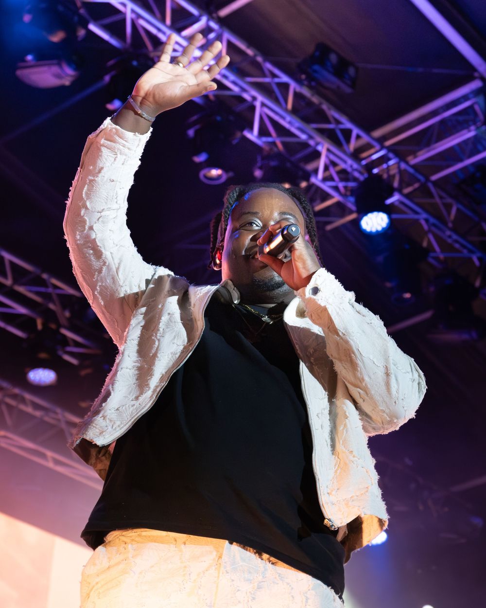 Performer singing on stage with one hand raised, wearing a white jacket and black top under colorful stage lights.