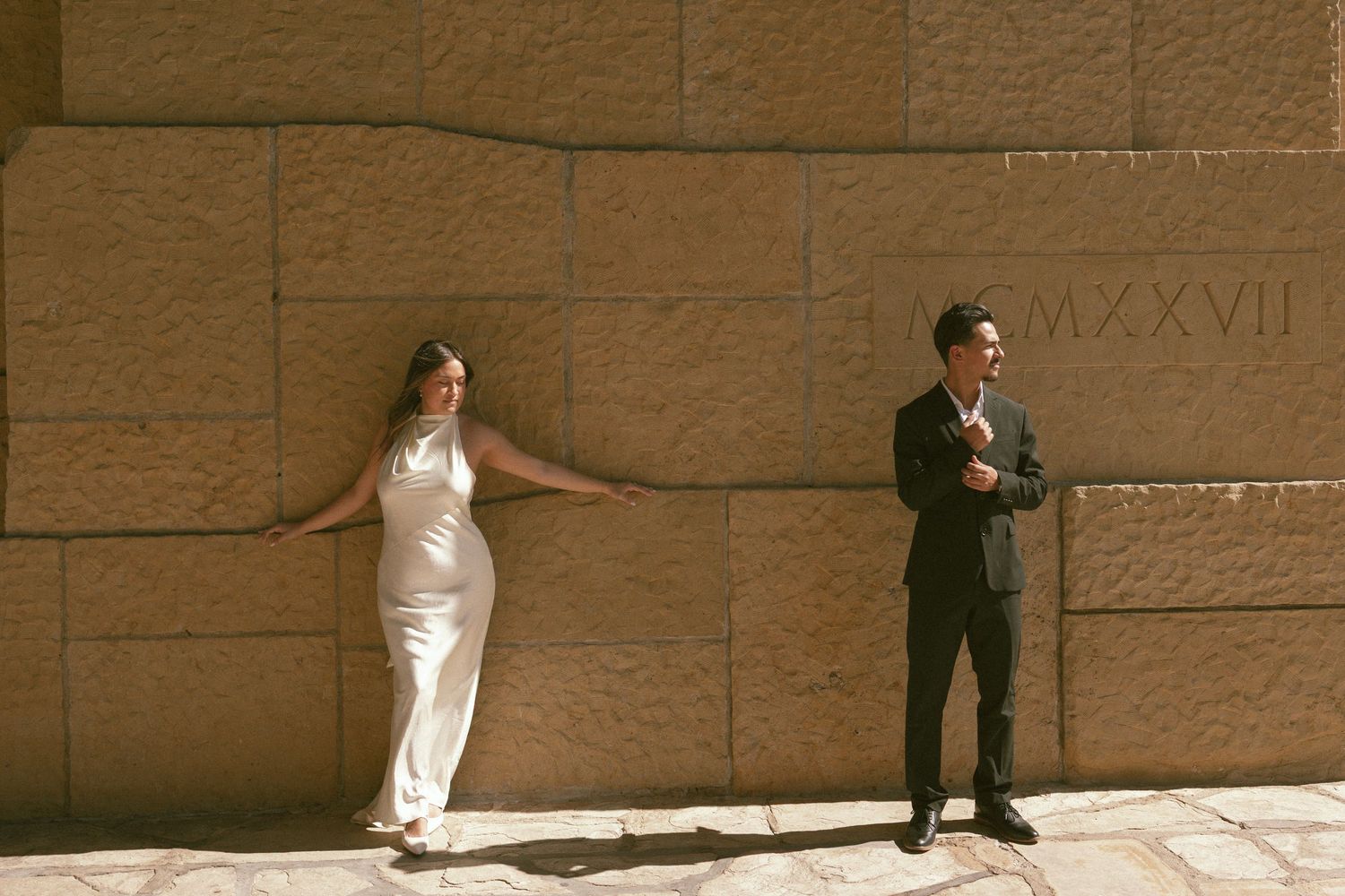 Couple posing by a stone wall with Roman numerals, woman in white dress and man in dark suit.