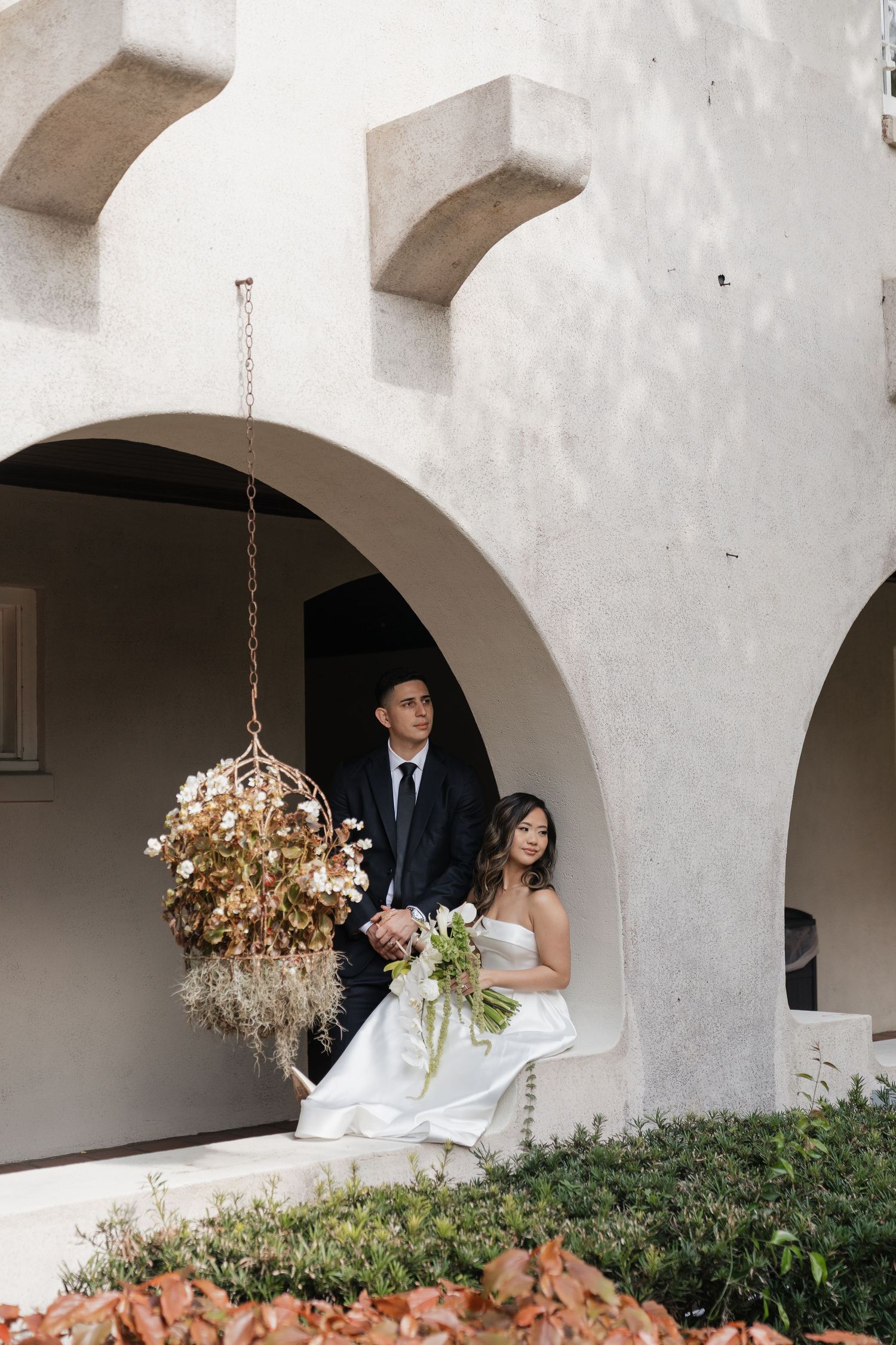Bride and groom pose under stone arches with hanging floral arrangement at elegant outdoor wedding venue.