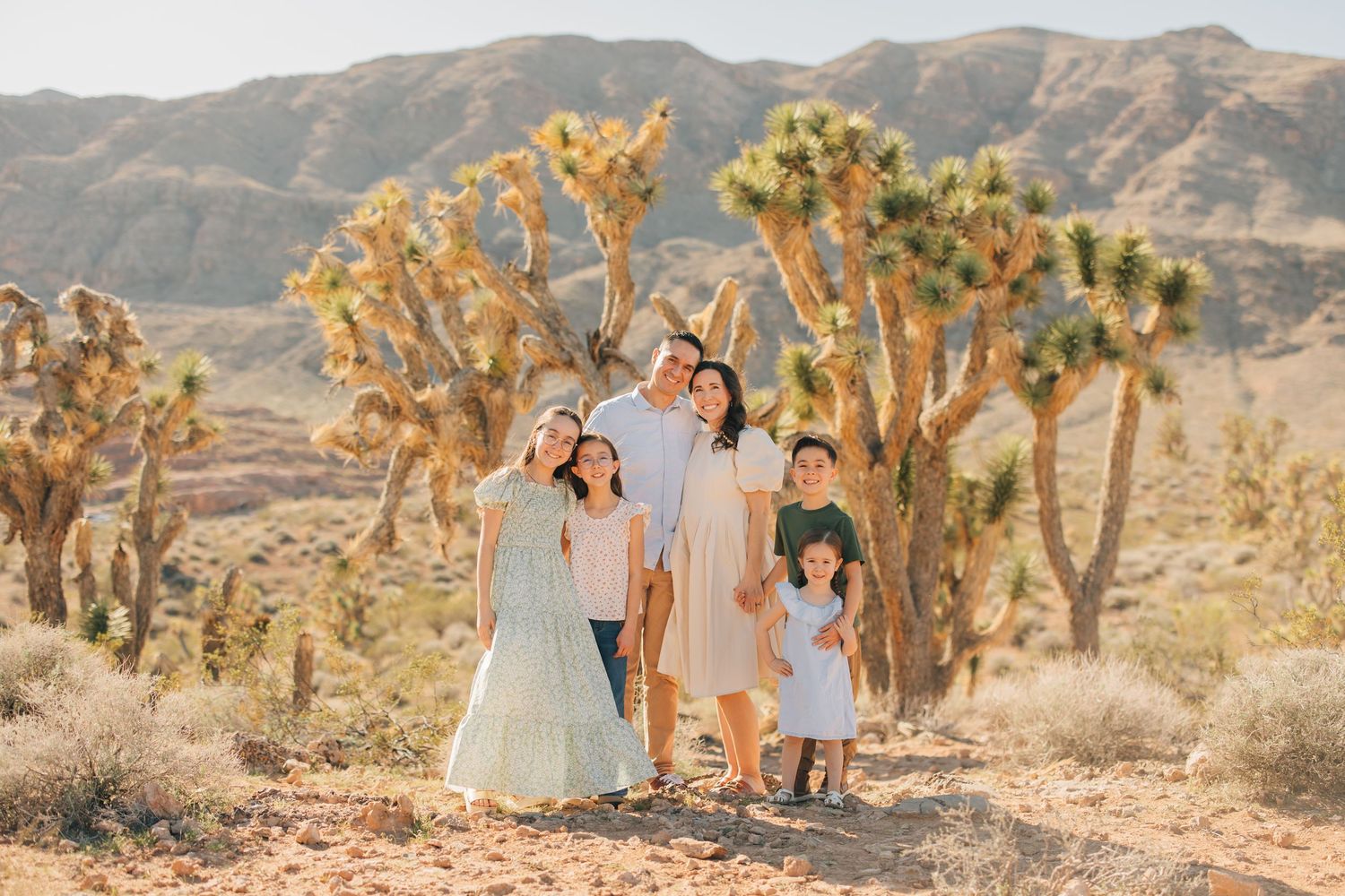 Family of six poses among Joshua trees in a sunlit desert landscape with mountains in the background.