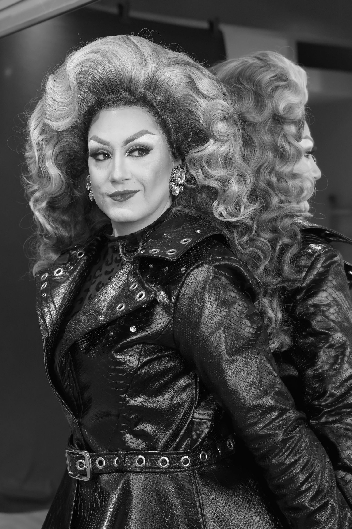 Black and white portrait of a drag performer in voluminous blonde wig and black leather outfit, posing confidently.