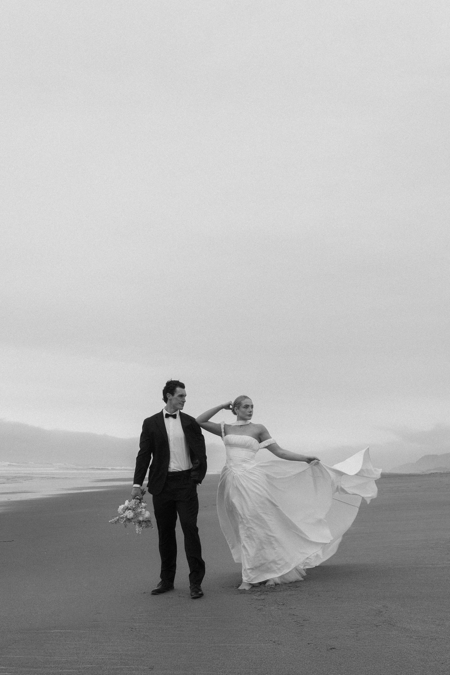 Black and white photo of a bride and groom walking on a beach, her gown flowing in the wind.