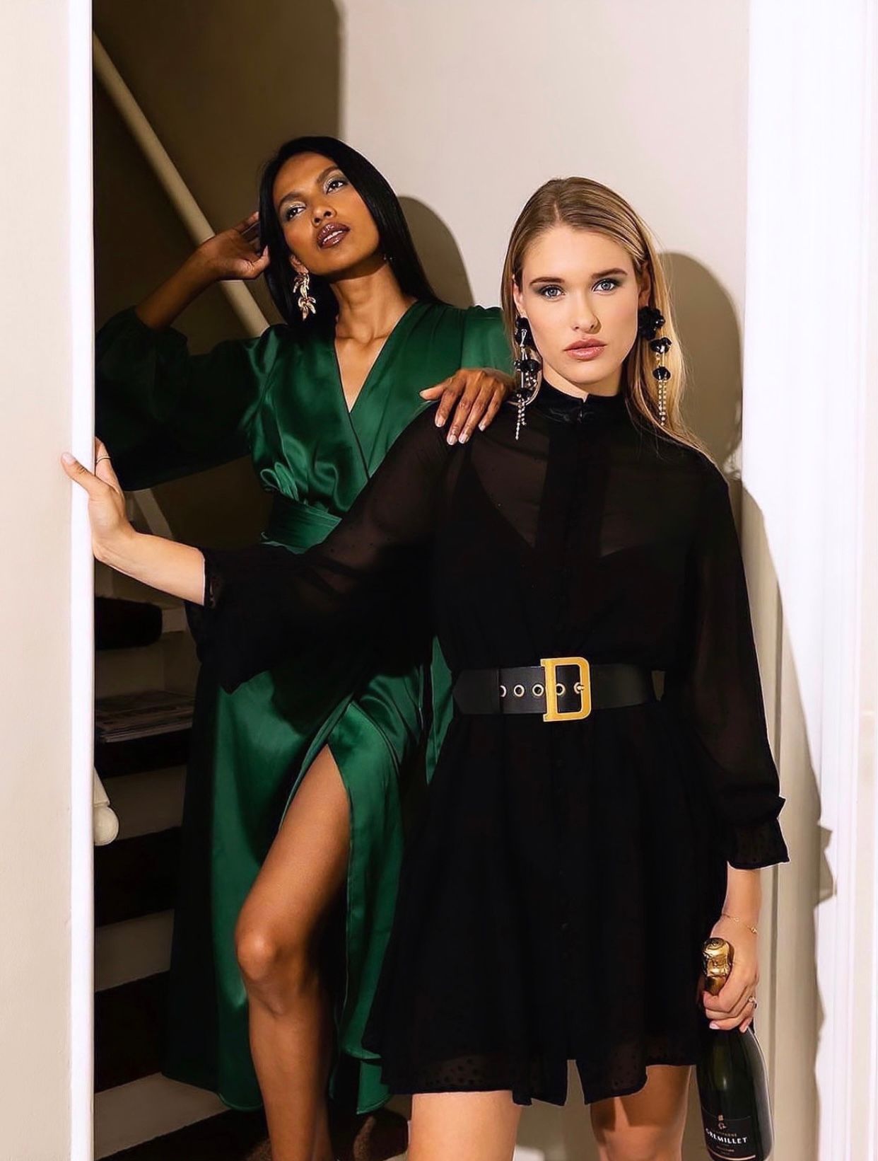 Two models pose on stairs, one in emerald satin wrap dress, other in black chiffon mini dress with gold belt.