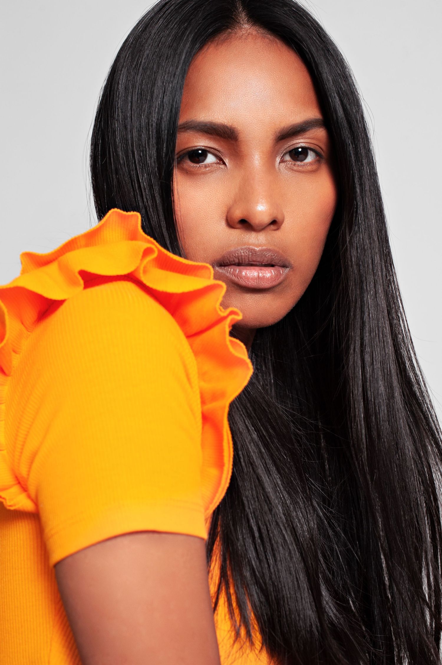 Woman with long black hair wearing a bright orange ruffled short-sleeve top, posing against a gray background.