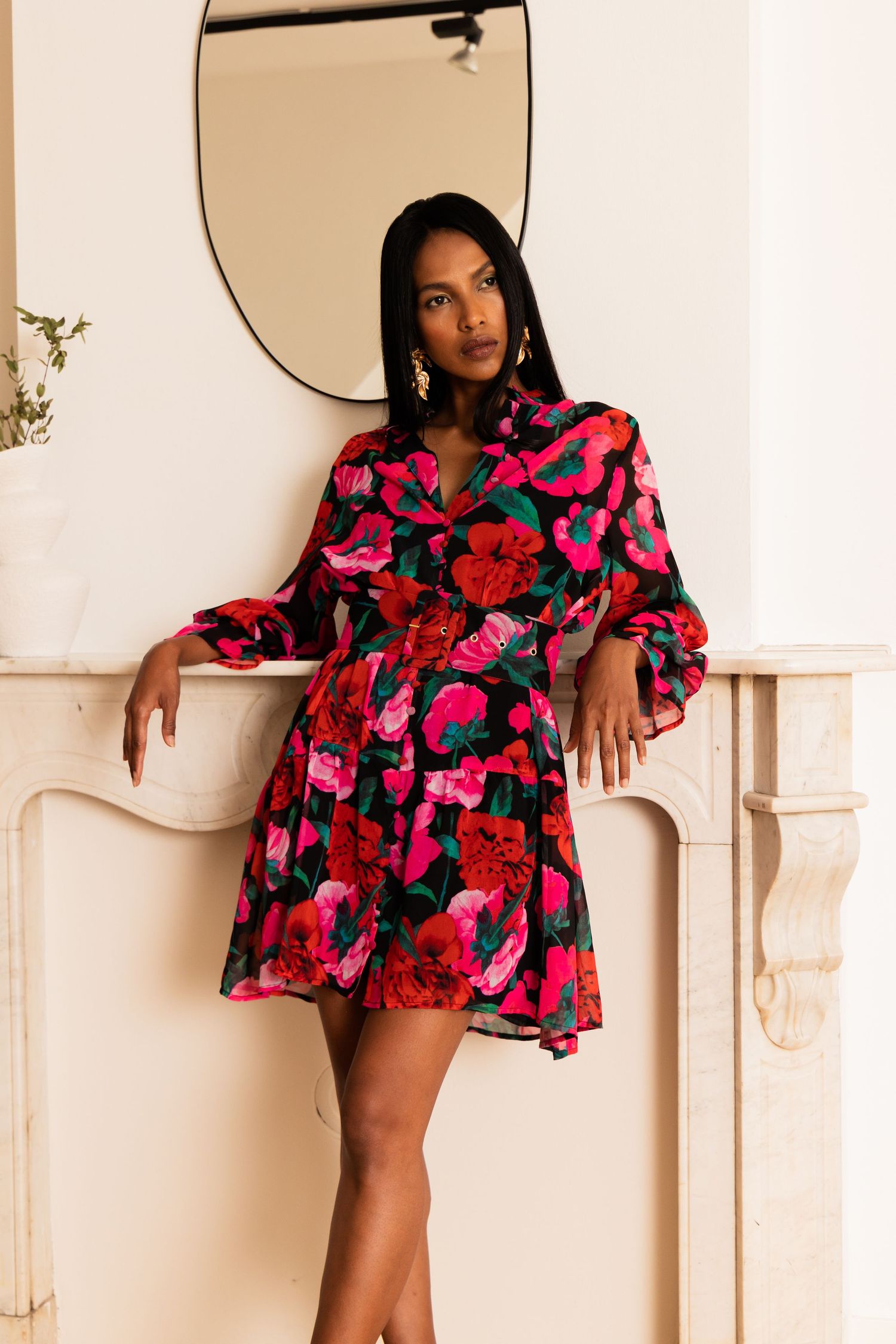 Woman in black floral mini dress with red and pink blooms, long sleeves, posing by a white fireplace mantel.