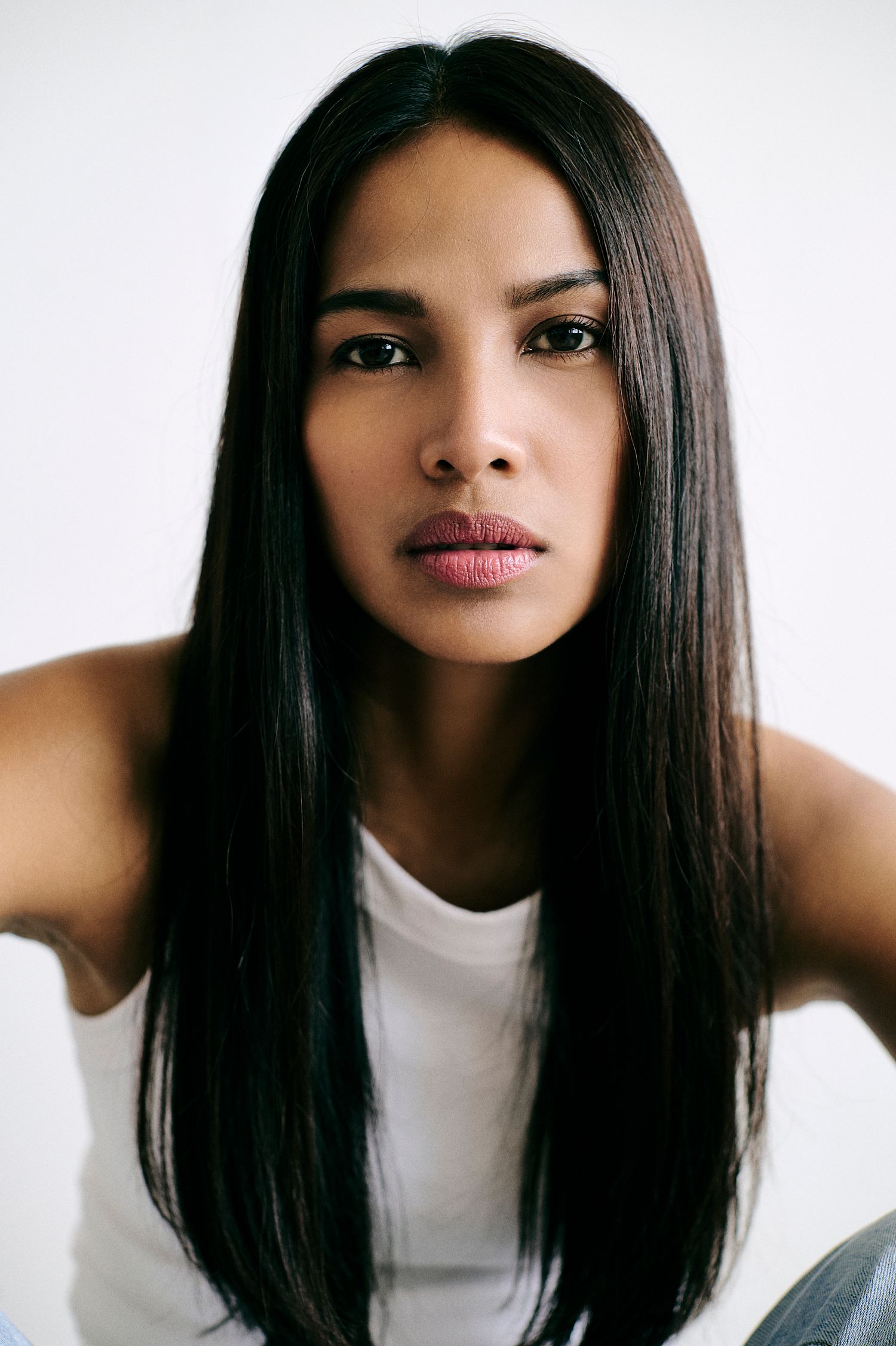 Close-up portrait of woman with long straight black hair wearing white tank top against white background.