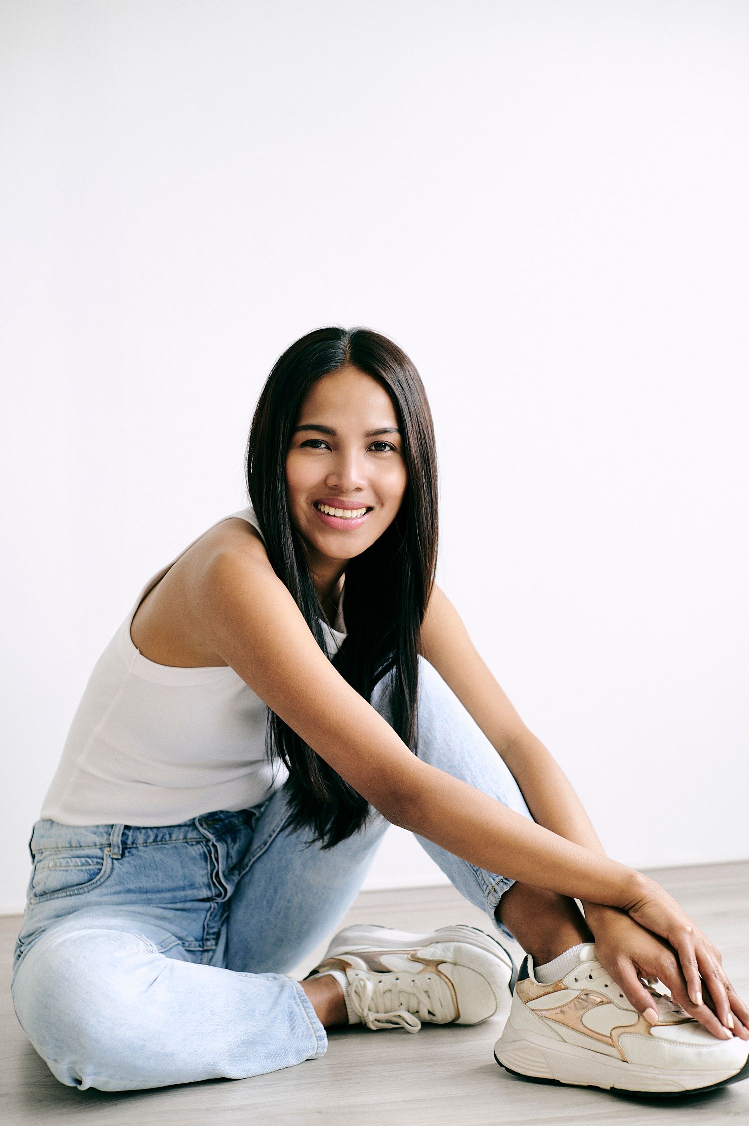 Young woman with long dark hair smiling, sitting on floor in white tank top, blue jeans, and white sneakers.