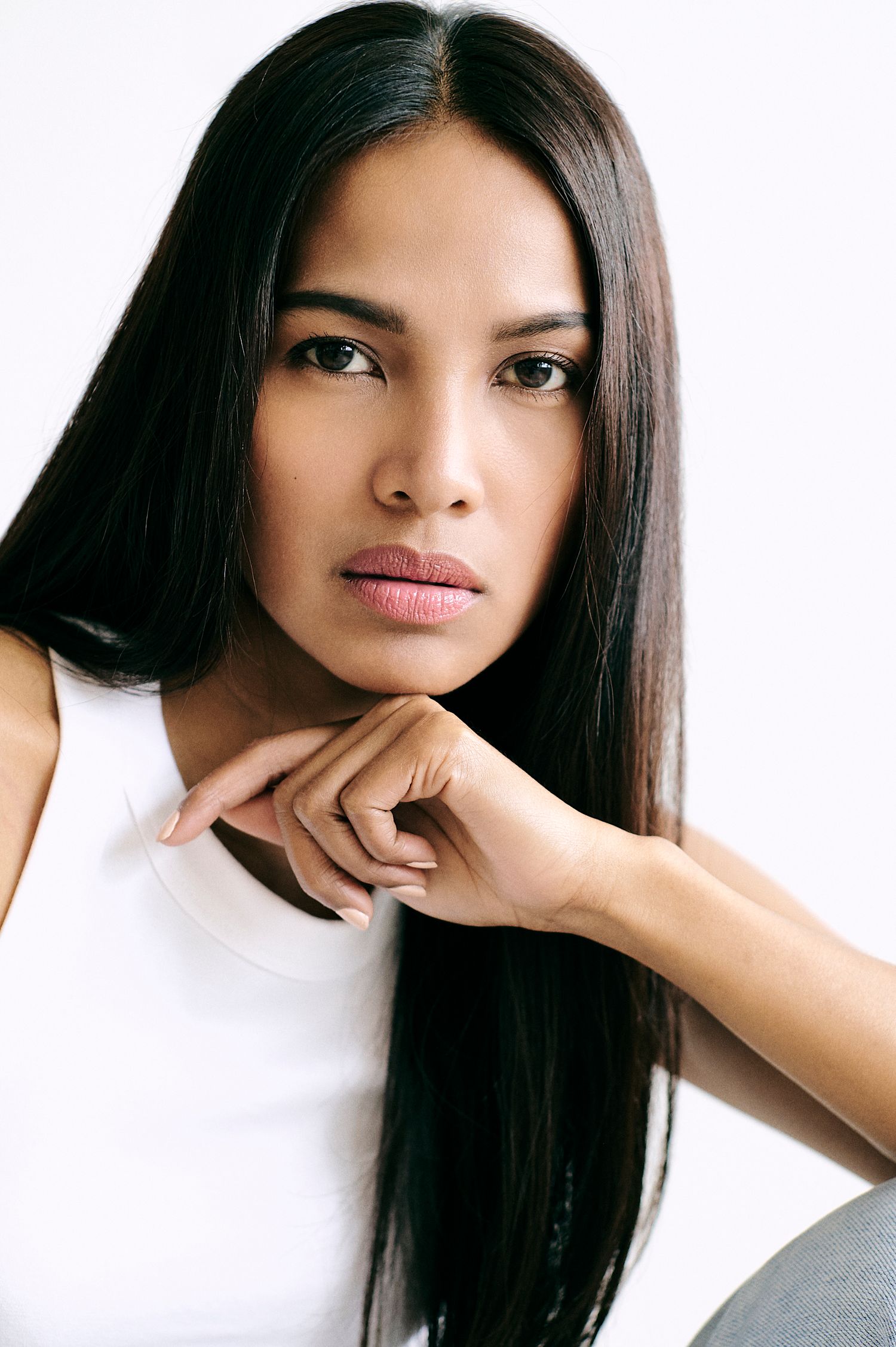 Young woman with long straight black hair poses thoughtfully with hand under chin, wearing a white sleeveless top.